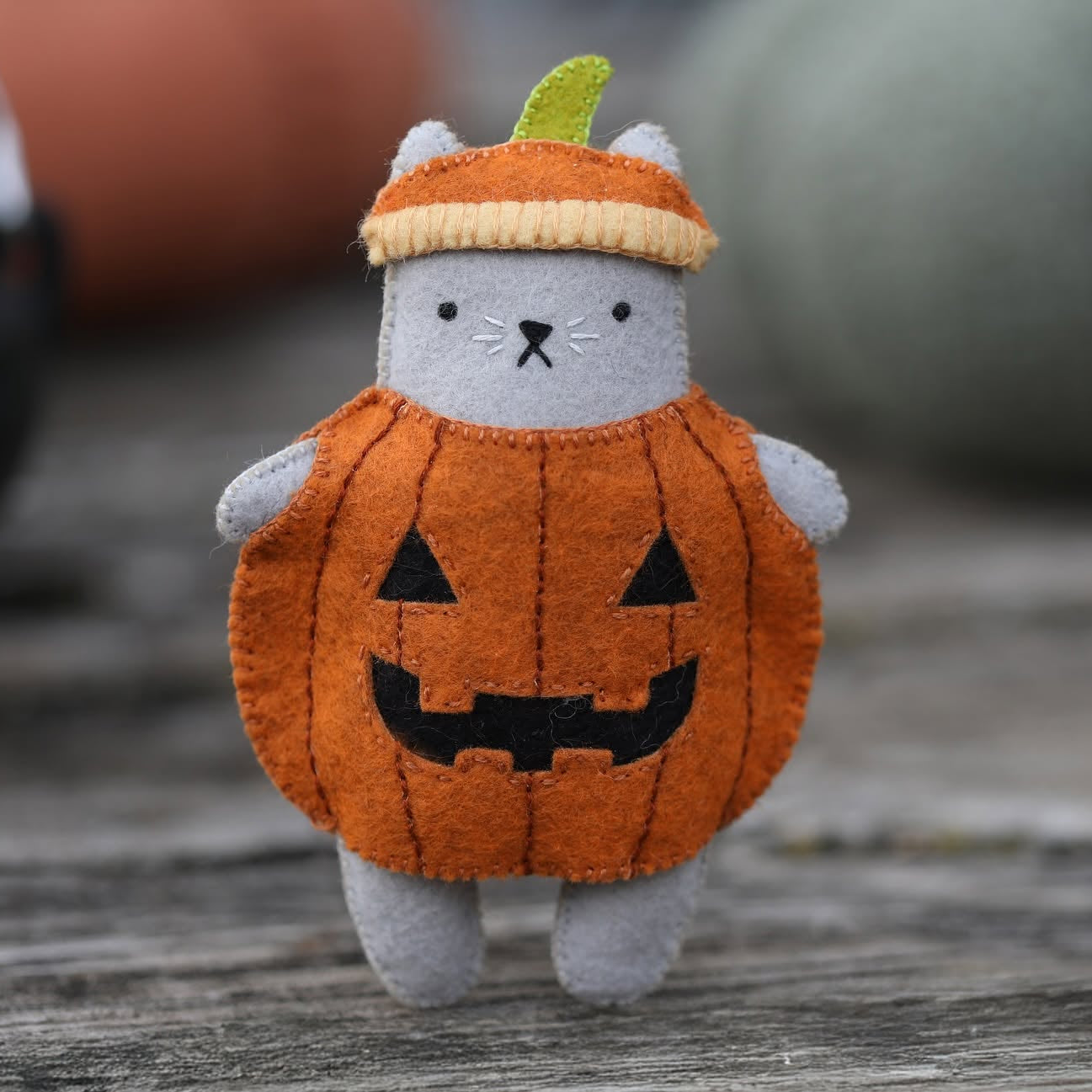 A small plush cat toy dressed in an orange pumpkin costume with a green leaf on top, standing on a weathered wooden surface with a softly blurred outdoor background.