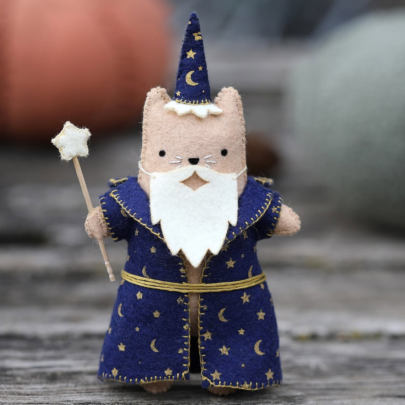 A small felt wizard cat figurine wearing a navy blue robe and pointed hat with gold stars and moons, holding a star wand, photographed on a rustic wooden surface with a softly blurred background.