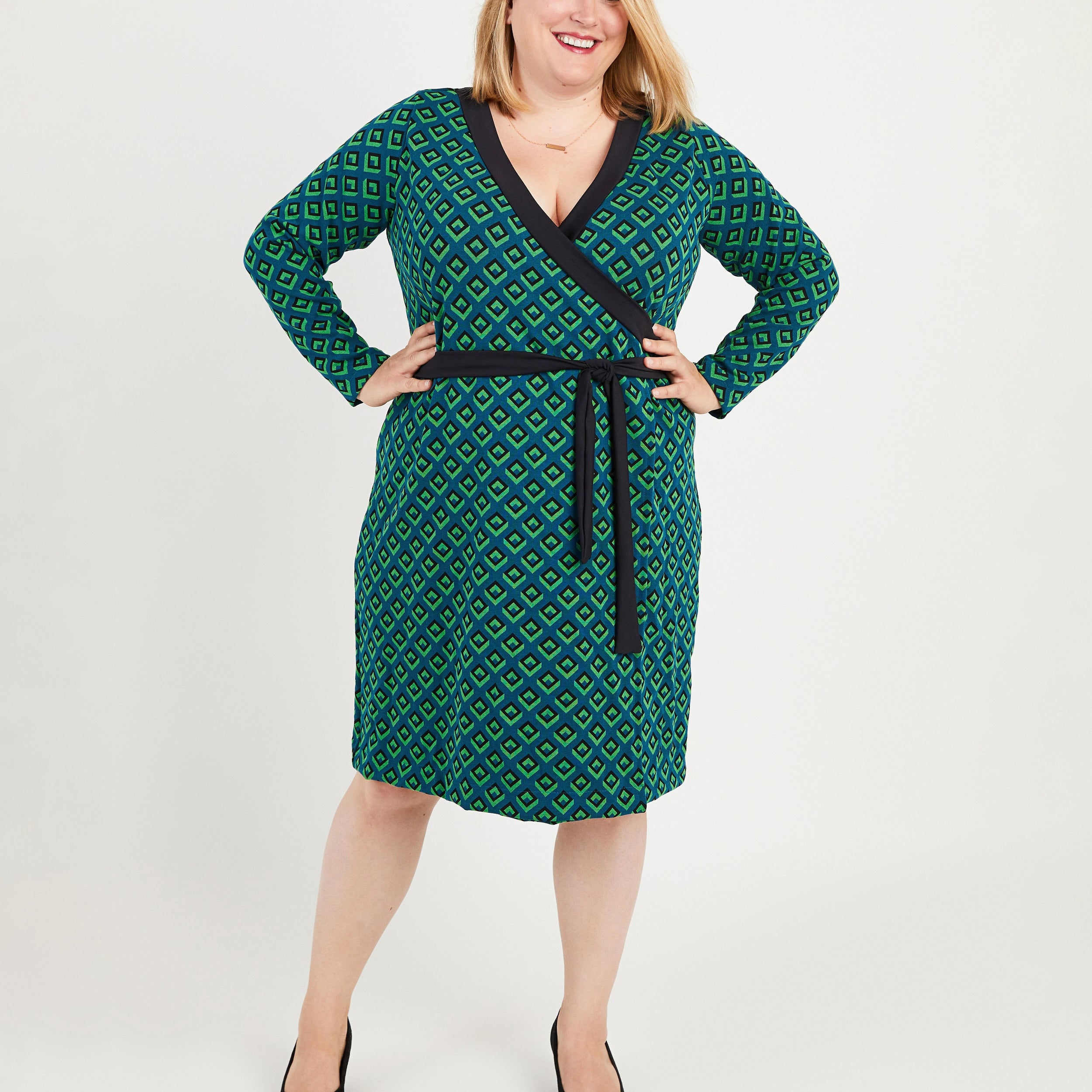 Woman wearing a green patterned dress with a belt on a white background. This dress is the Appleton Wrap Dress sewing pattern from Cashmerette.