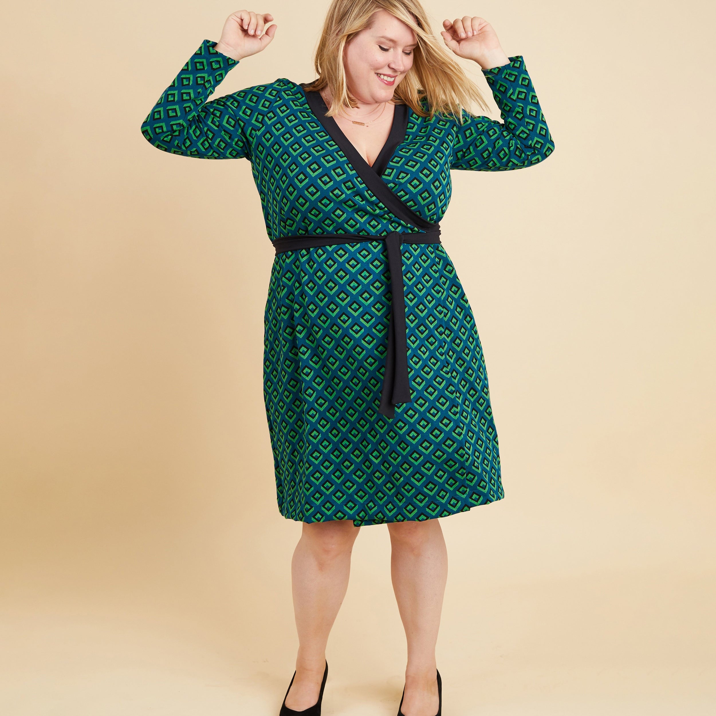 Woman wearing a green patterned dress with a belt on a beige background. This dress is the Appleton Wrap Dress sewing pattern from Cashmerette.