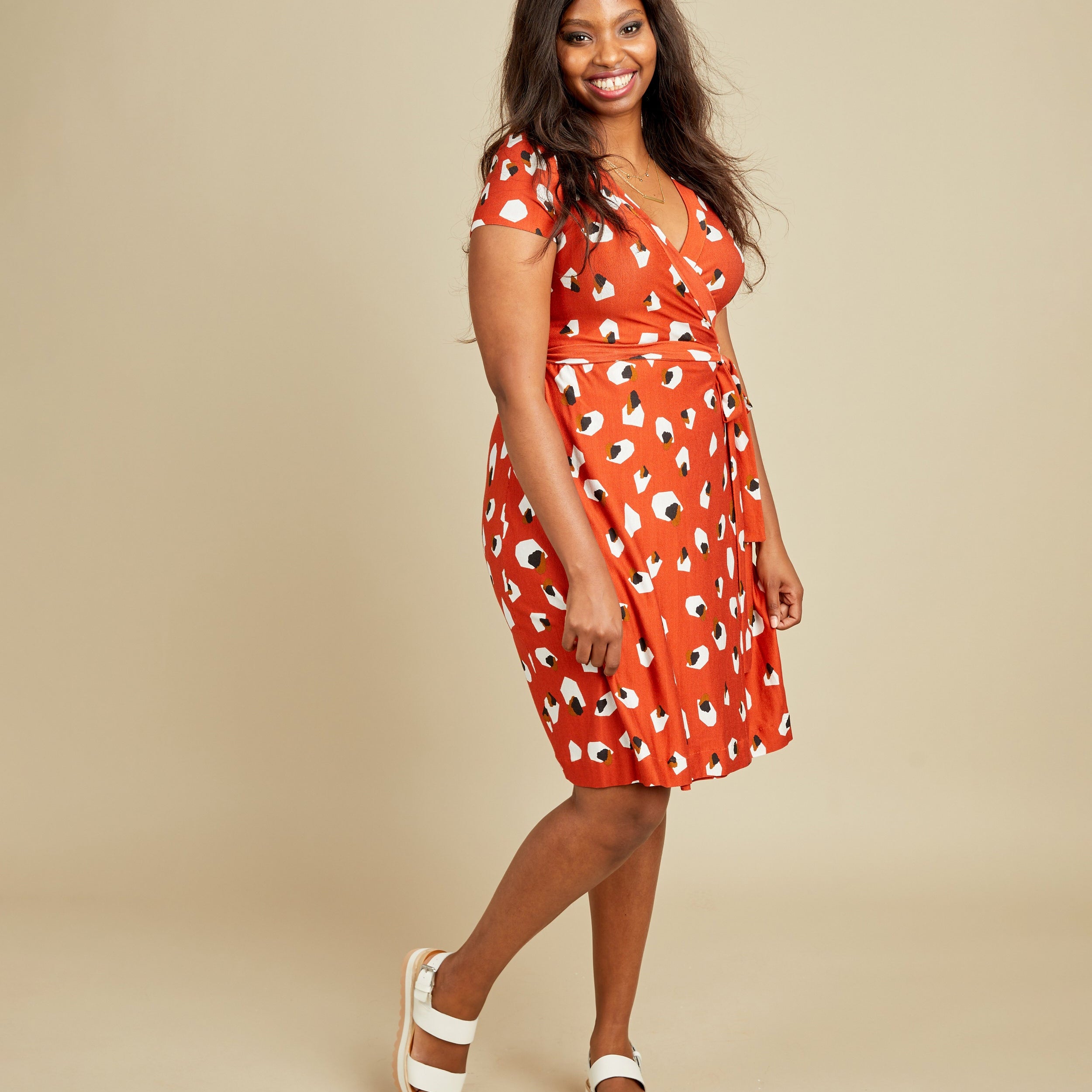 Woman wearing a red dress with white patterns on a beige background. This dress is the Appleton Wrap Dress sewing pattern from Cashmerette.