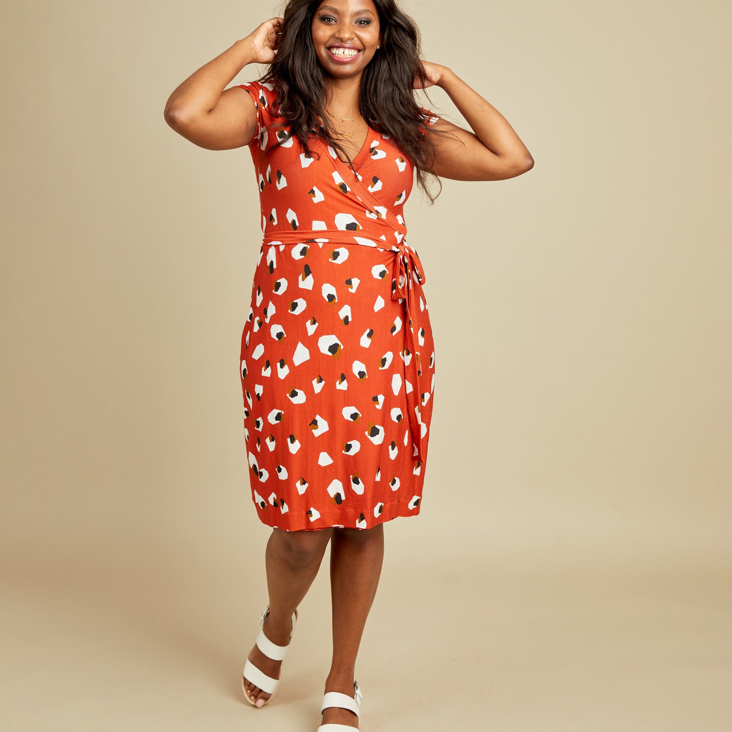 Woman wearing a red dress with a pattern on a beige background. This dress is the Appleton Wrap Dress sewing pattern from Cashmerette.