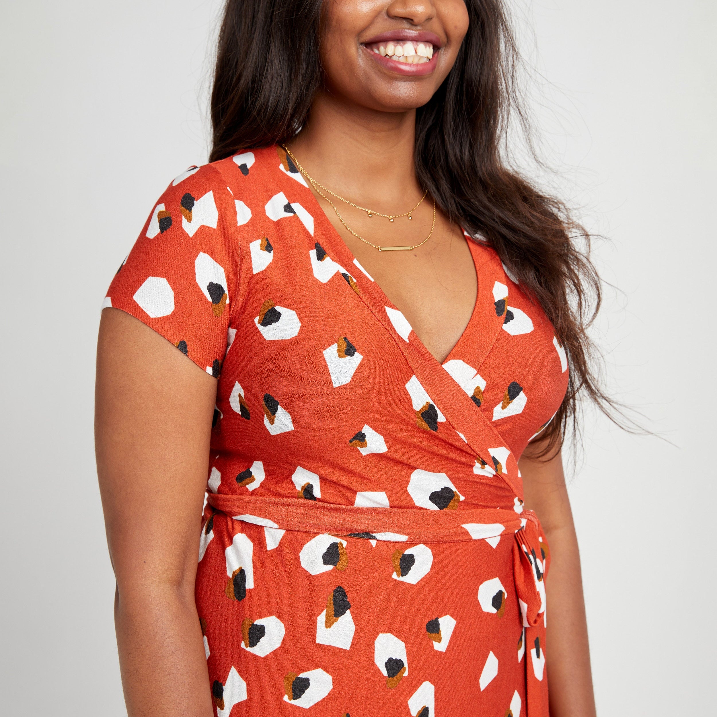 Woman wearing an orange dress with a pattern of white and black shapes on a plain background. This dress is the Appleton Wrap Dress sewing pattern from Cashmerette.
