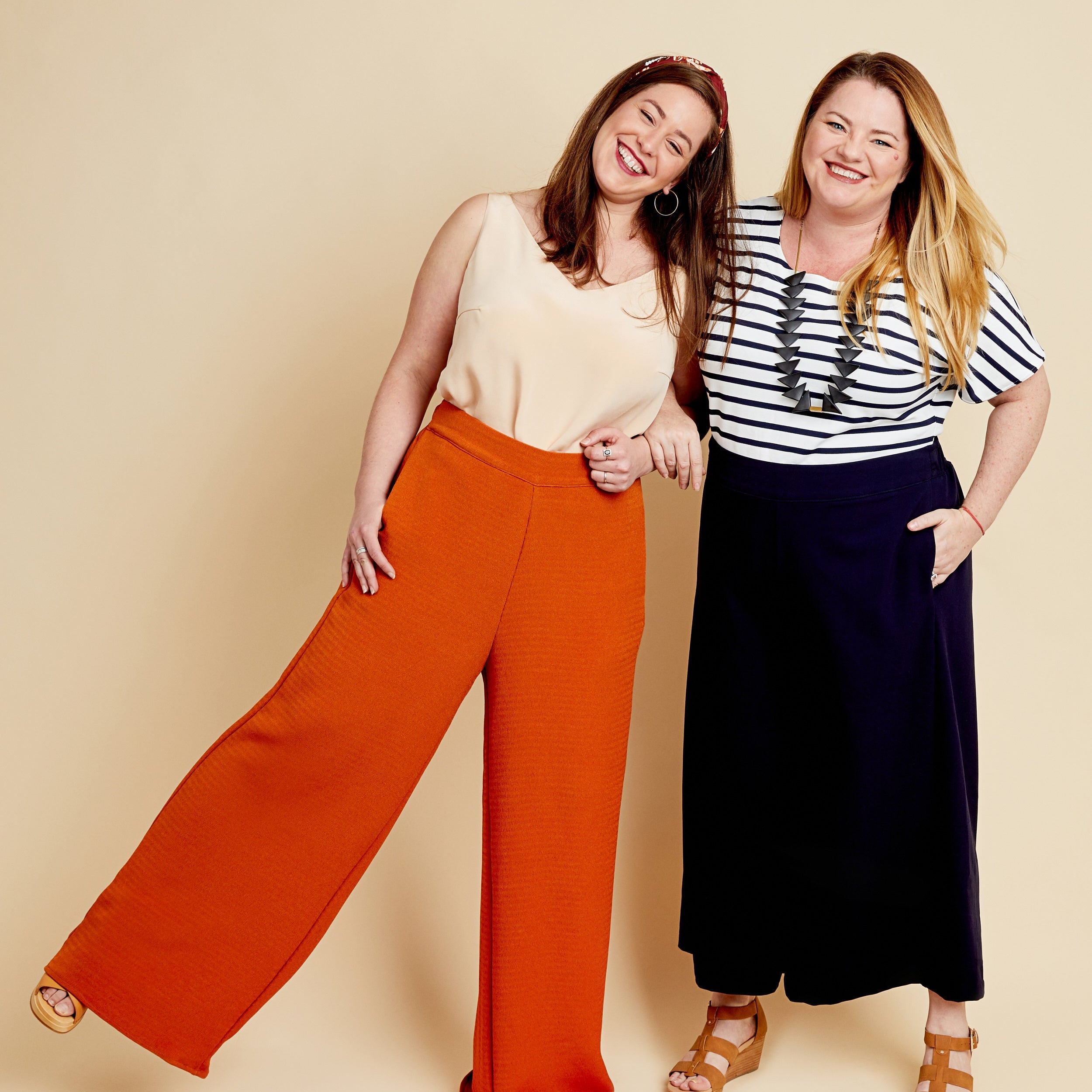 Two women standing side by side against a beige background, one wearing orange pants and a beige top, the other in a striped shirt and navy pants. These pants are the Calder Pants & Shorts sewing pattern from Cashmerette.