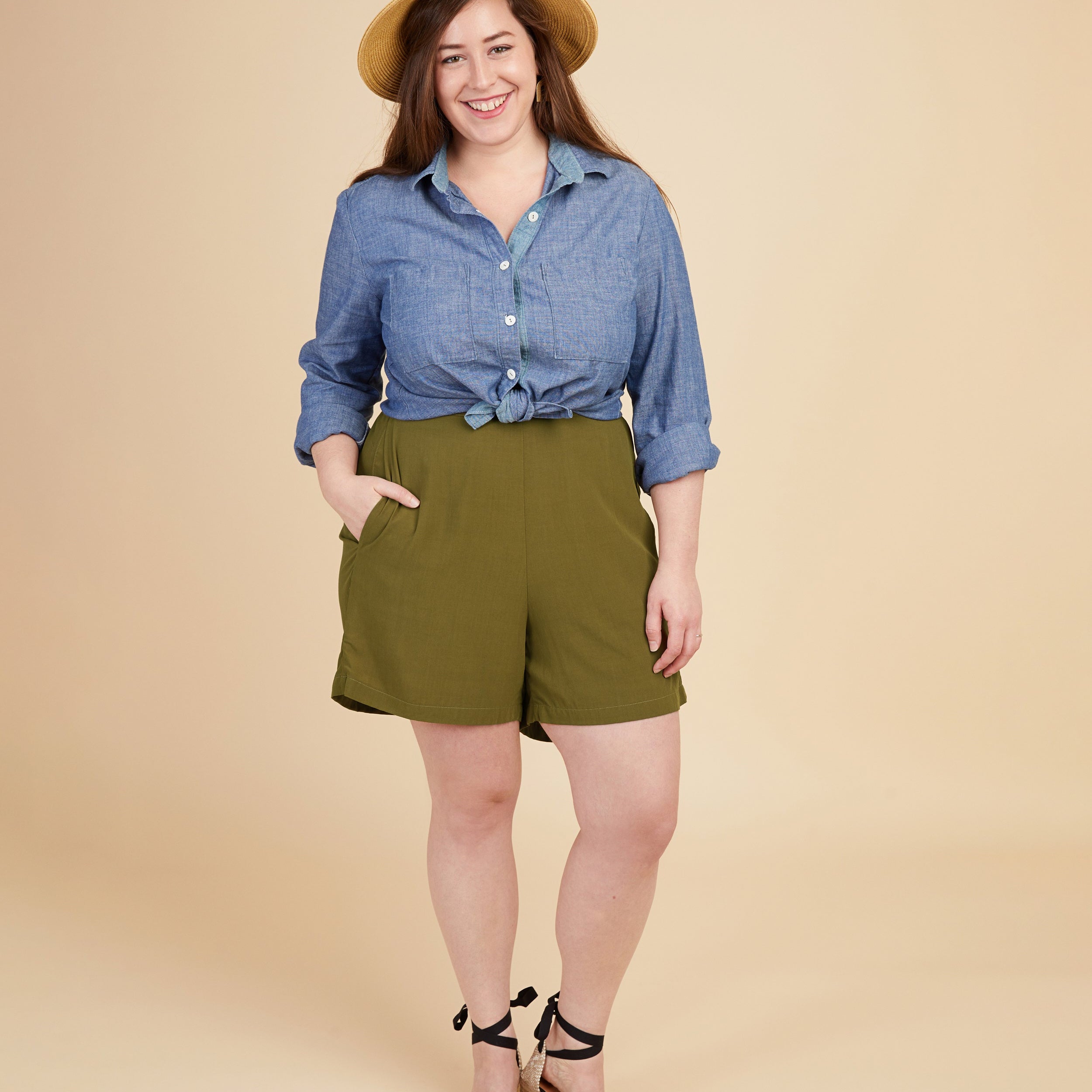 Woman wearing a blue shirt and green shorts with a beige hat on a beige background. These shorts are the Calder Pants & Shorts sewing pattern from Cashmerette.