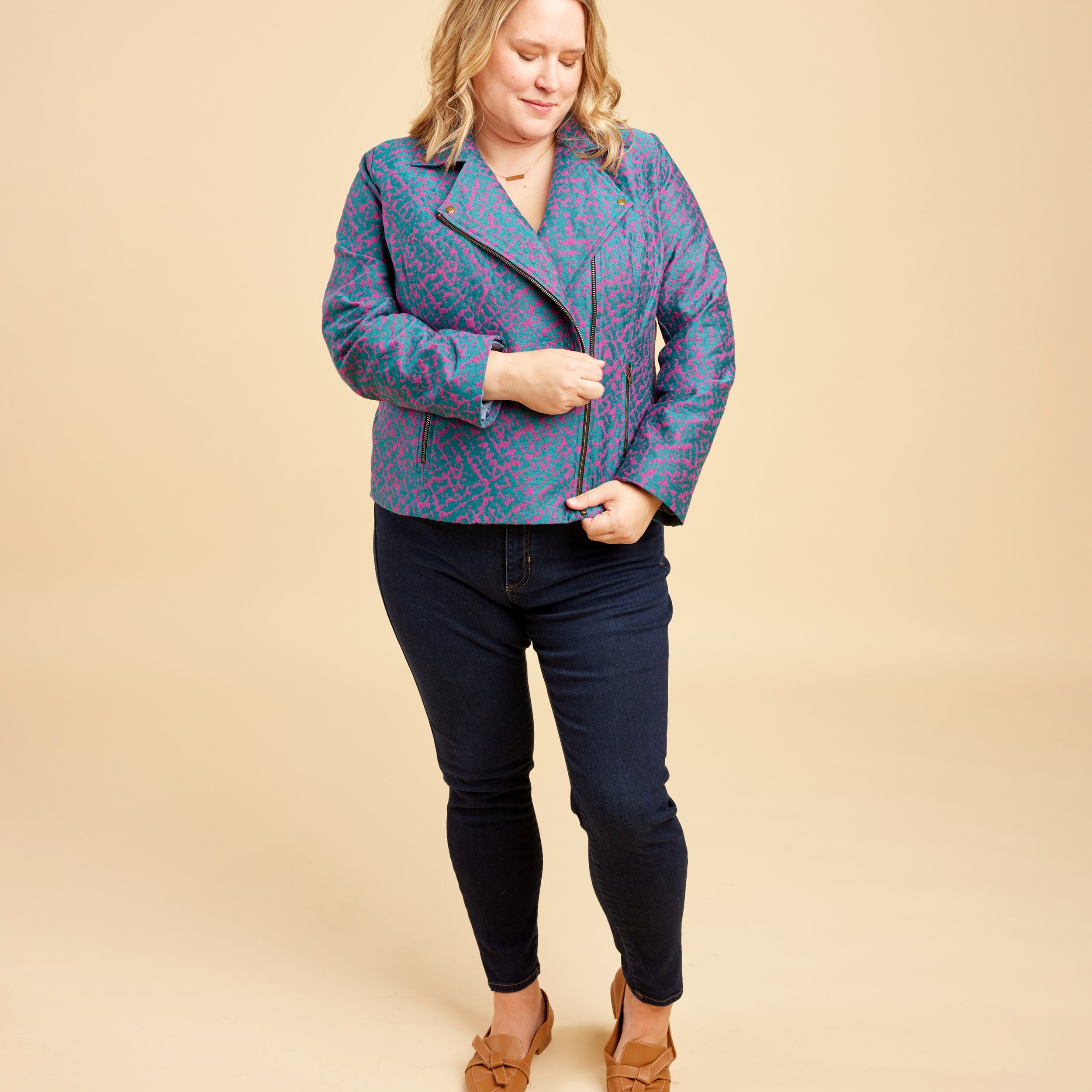 Woman wearing a blue patterned jacket and dark jeans on a beige background. This biker jacket is the Canton Moto Jacket sewing pattern from Cashmerette.