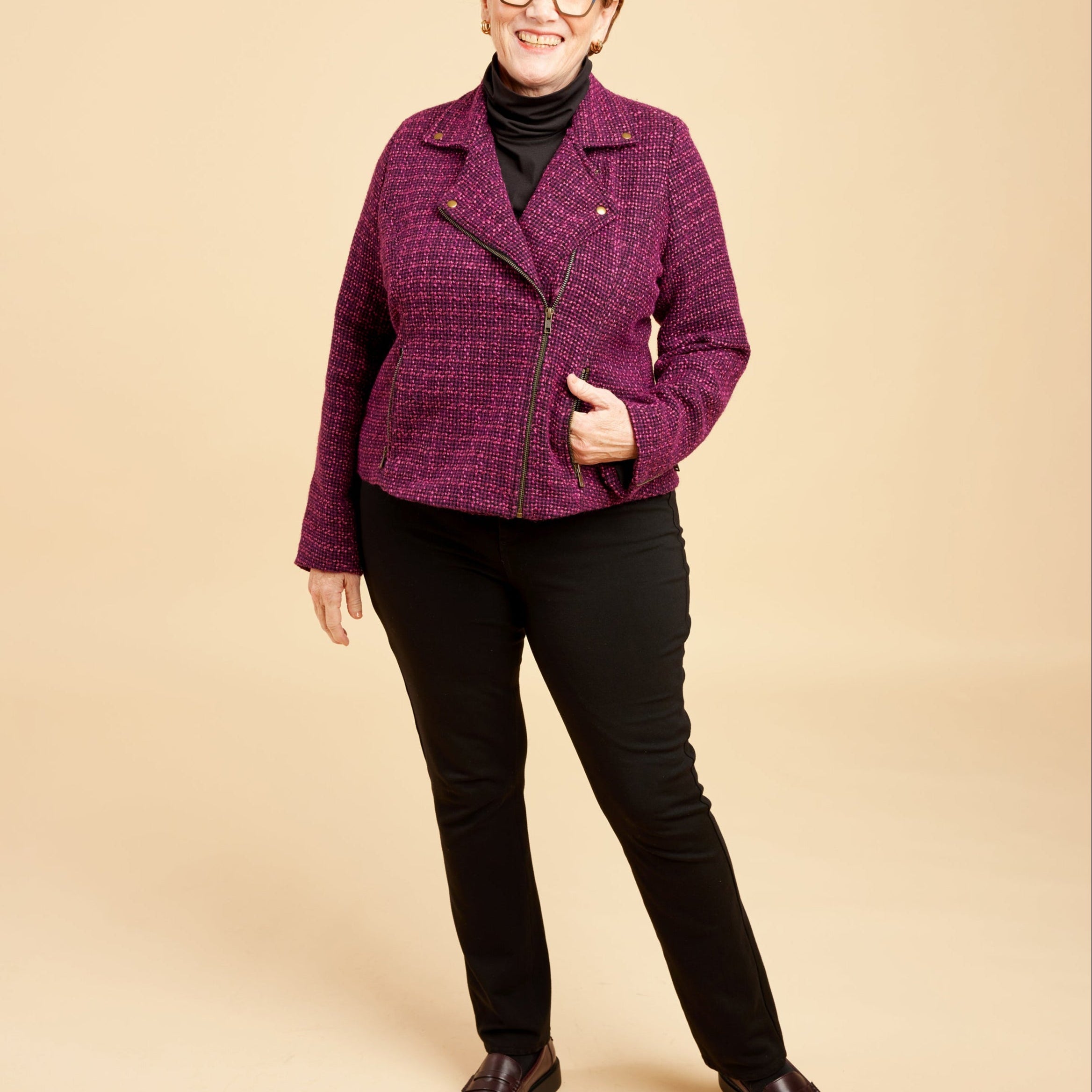 Woman wearing a purple jacket and black pants on a beige background. This biker jacket is the Canton Moto Jacket sewing pattern from Cashmerette.