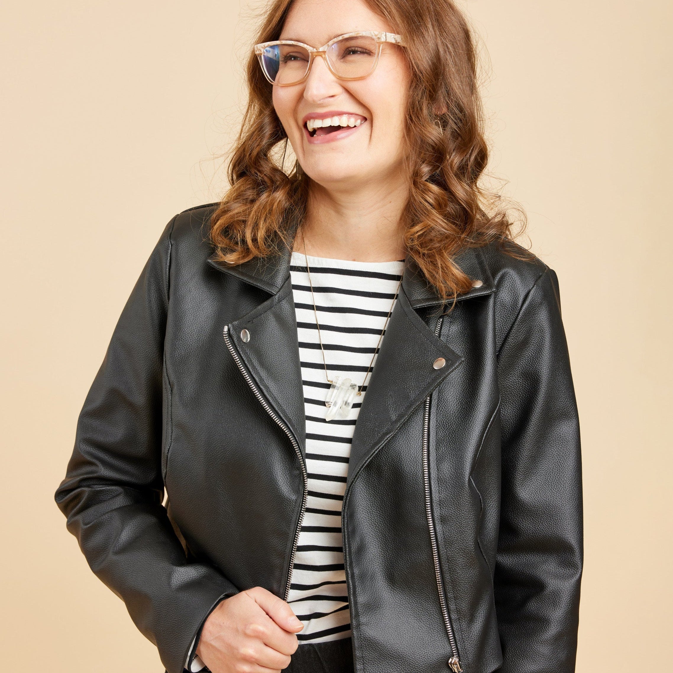 Woman wearing a black leather jacket over a striped shirt on a beige background. This biker jacket is the Canton Moto Jacket sewing pattern from Cashmerette.