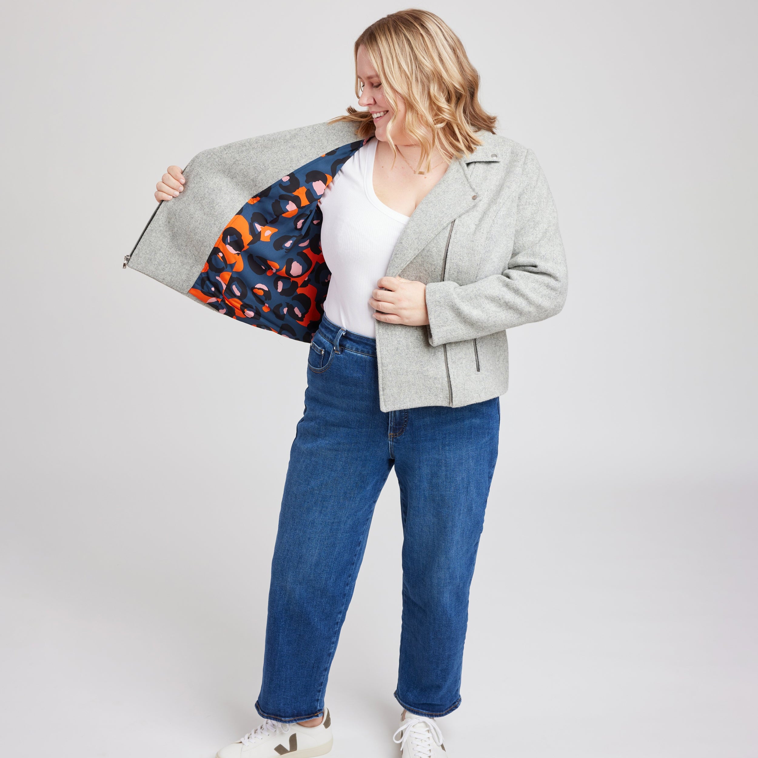 Person wearing a gray jacket with a colorful lining, white shirt, blue jeans, and white sneakers on a light gray background. This biker jacket is the Canton Moto Jacket sewing pattern from Cashmerette.