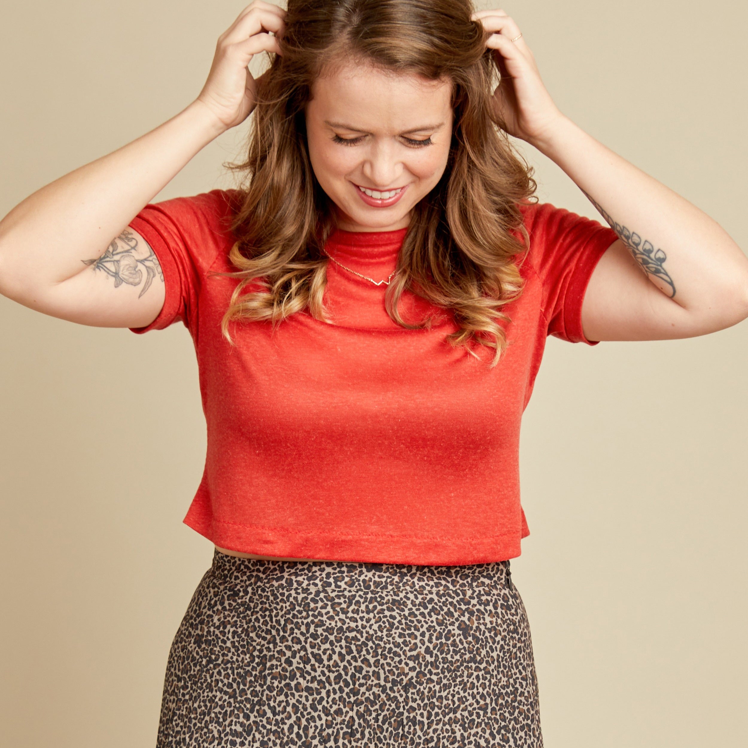 Woman wearing a red shirt and patterned skirt adjusting her hair against a beige background. This t-shirt is the Concord T-Shirt sewing pattern from Cashmerette.