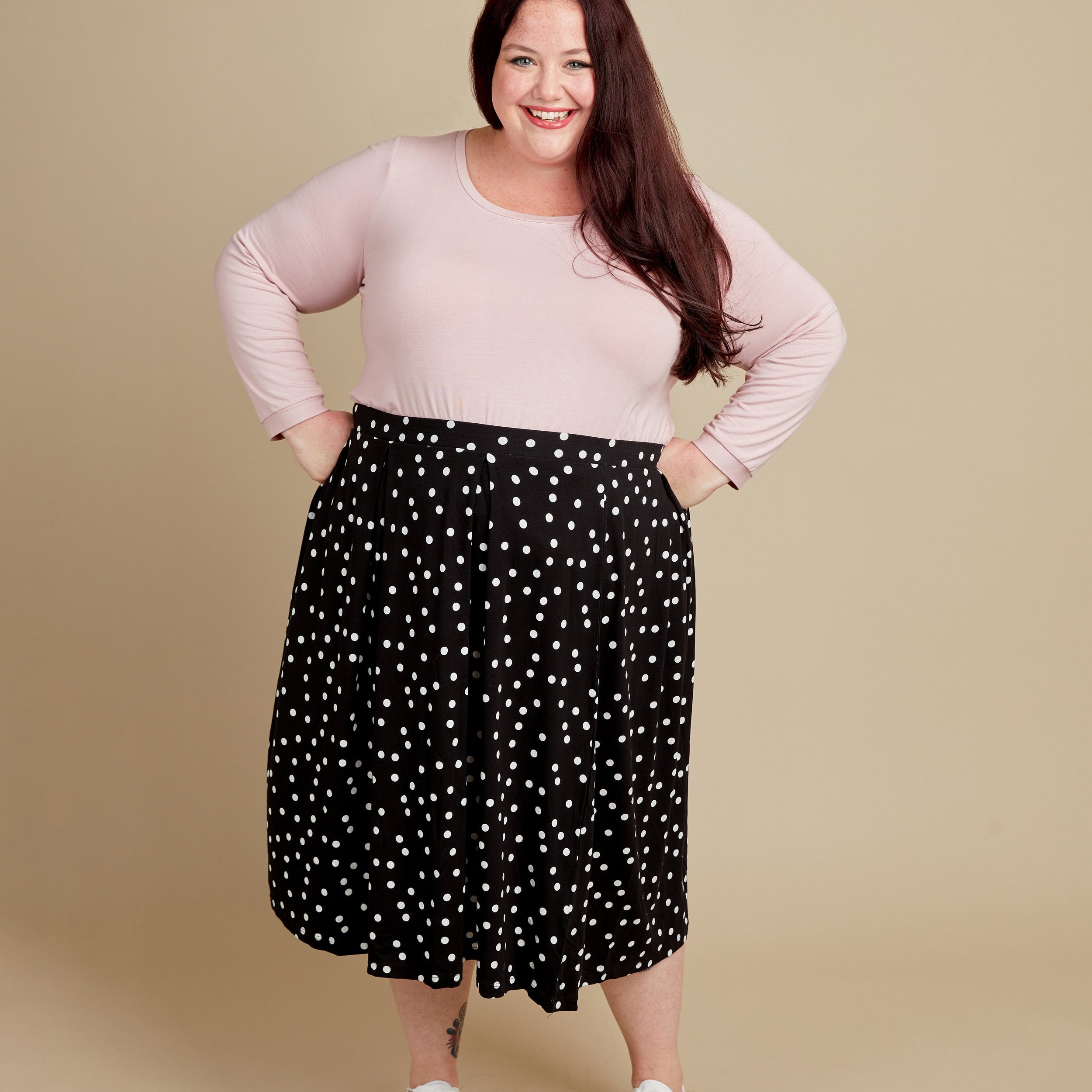 Woman wearing a pink top and black polka dot skirt on a beige background. This t-shirt is the Concord T-Shirt sewing pattern from Cashmerette.