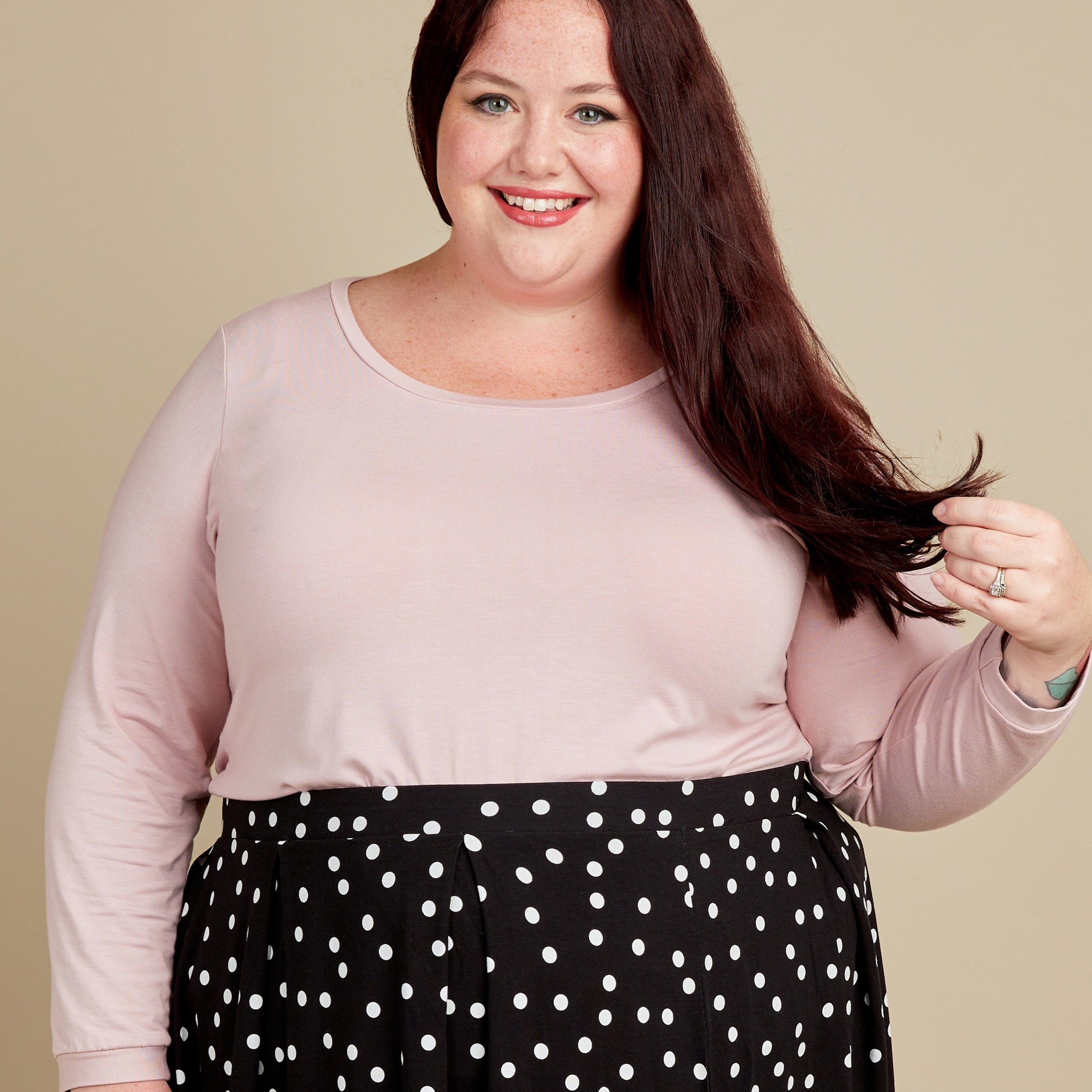 Woman wearing a light pink top and black polka dot pants against a beige background. This t-shirt is the Concord T-Shirt sewing pattern from Cashmerette.