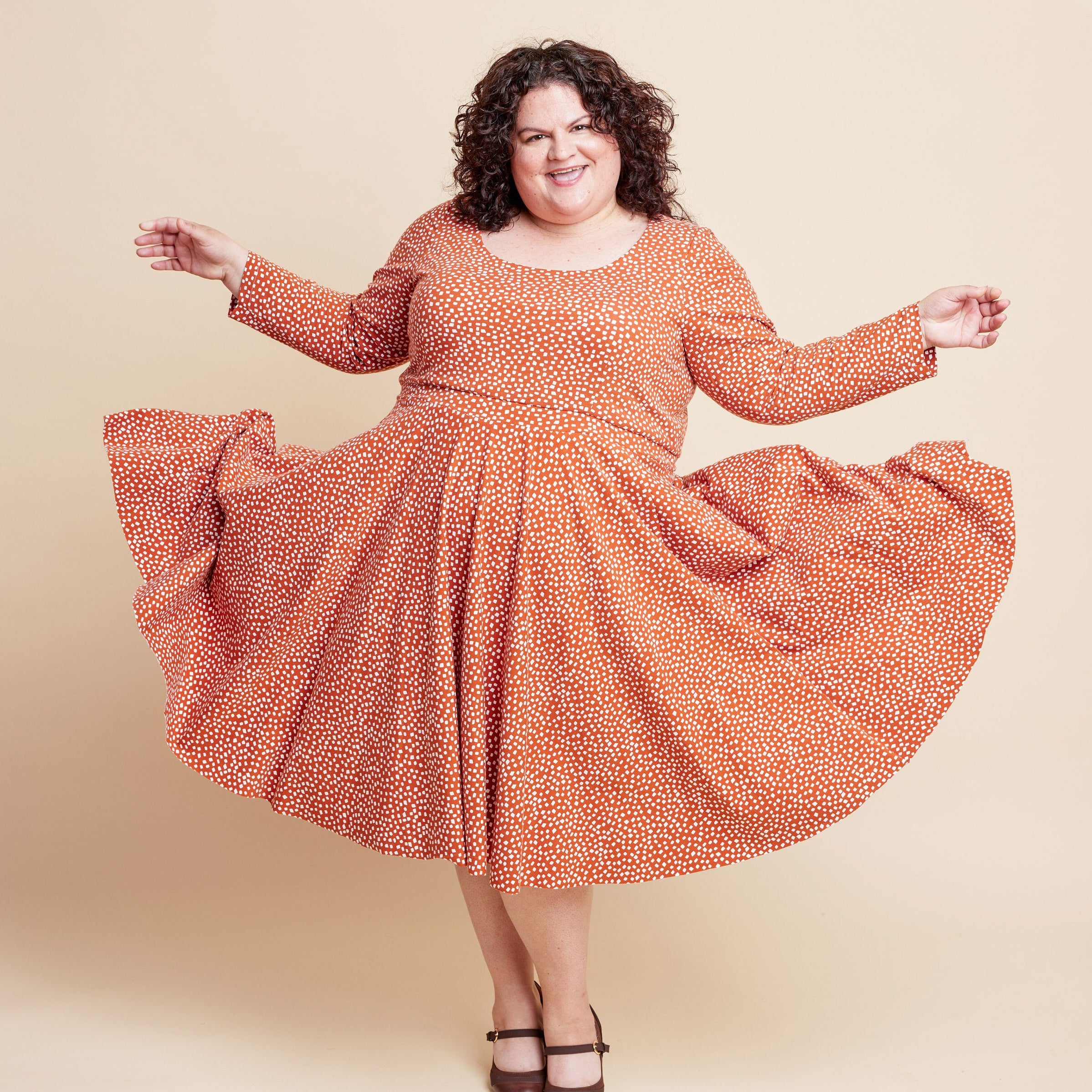 Woman wearing an orange patterned dress on a beige background. This is the Grafton Dress, Top & Skirt sewing pattern from Cashmerette.