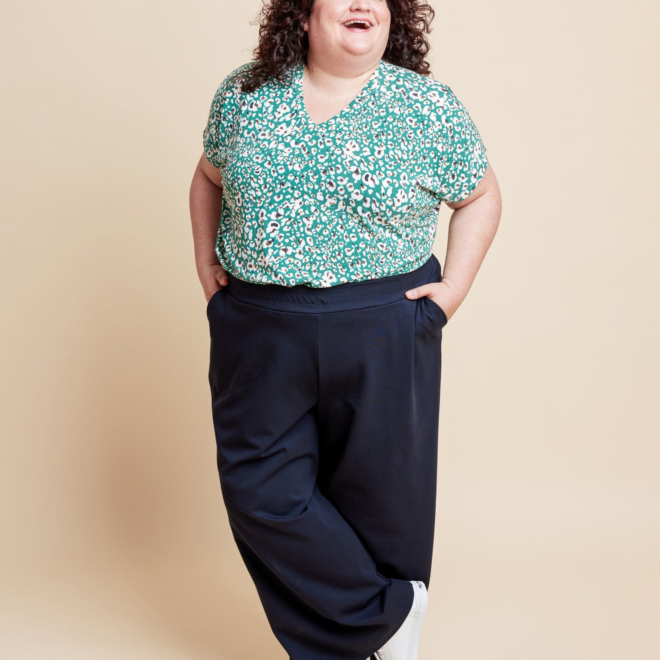 Woman wearing a floral top and dark pants on a beige background. This is the Grafton Dress, Top & Skirt sewing pattern from Cashmerette.
