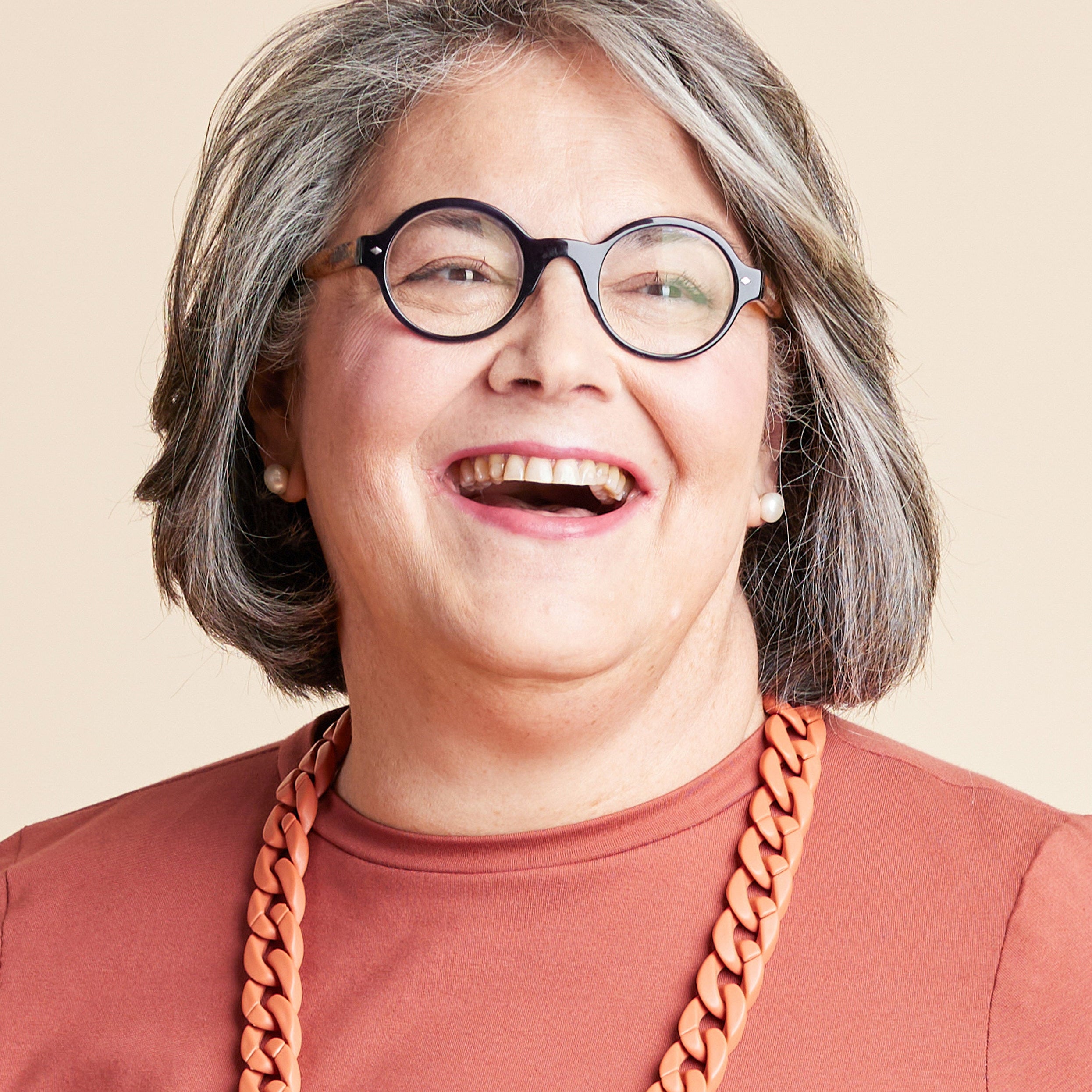 Person with gray hair wearing glasses and a brown shirt with a large orange necklace on a beige background. This is the Grafton Dress, Top & Skirt sewing pattern from Cashmerette.
