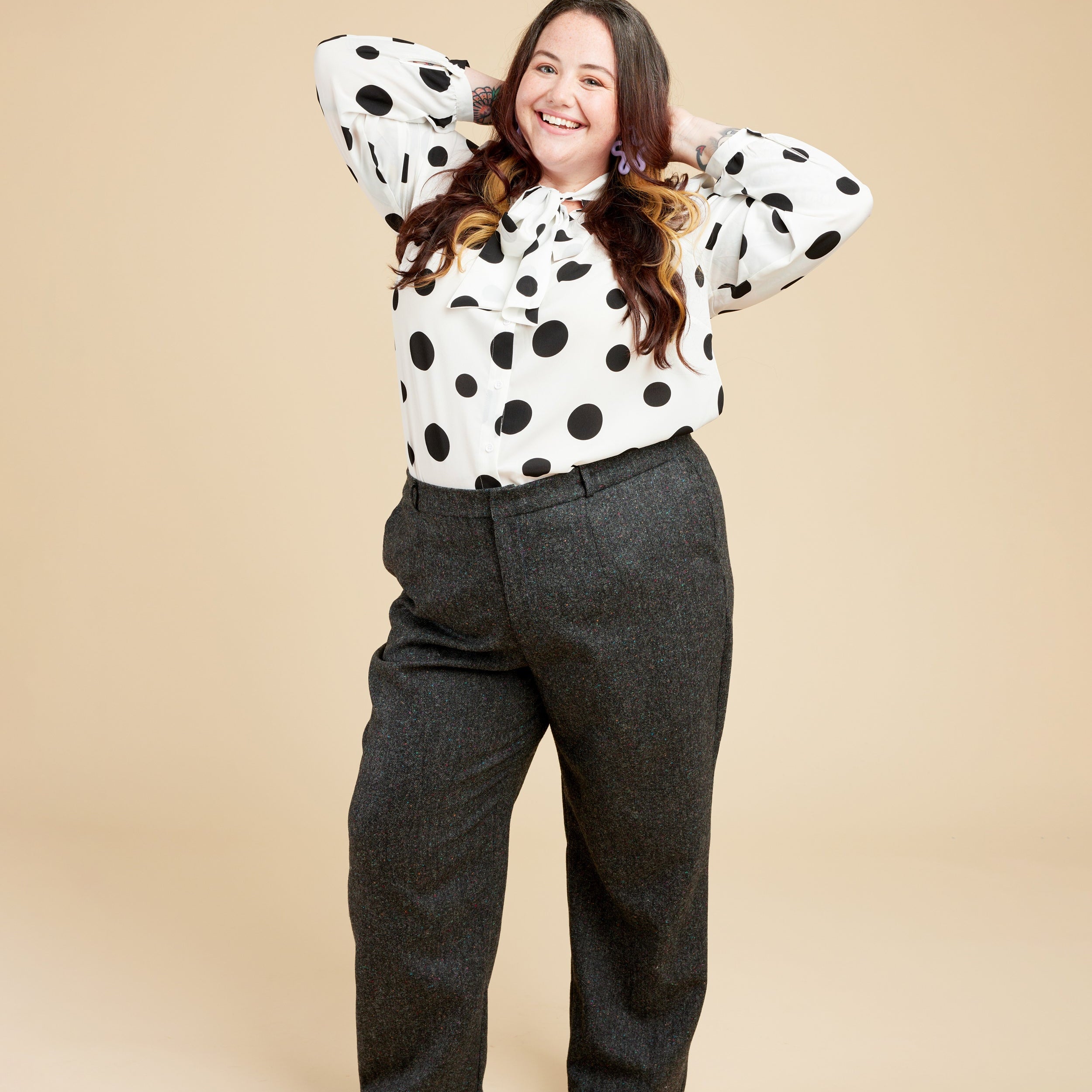 Person wearing a polka dot blouse and gray pants on a beige background. These pants are the Meriam Trousers sewing pattern from Cashmerette.