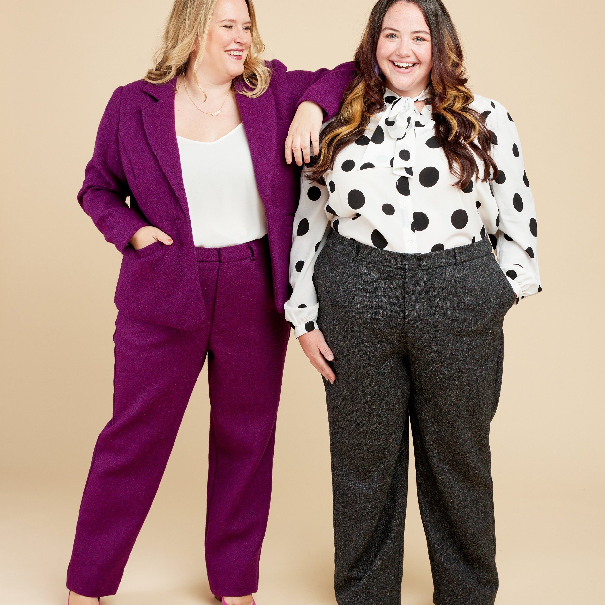 Two women posing together against a beige background, one in a purple suit and the other in a polka dot blouse and gray pants.. These pants are the Meriam Trousers sewing pattern from Cashmerette.