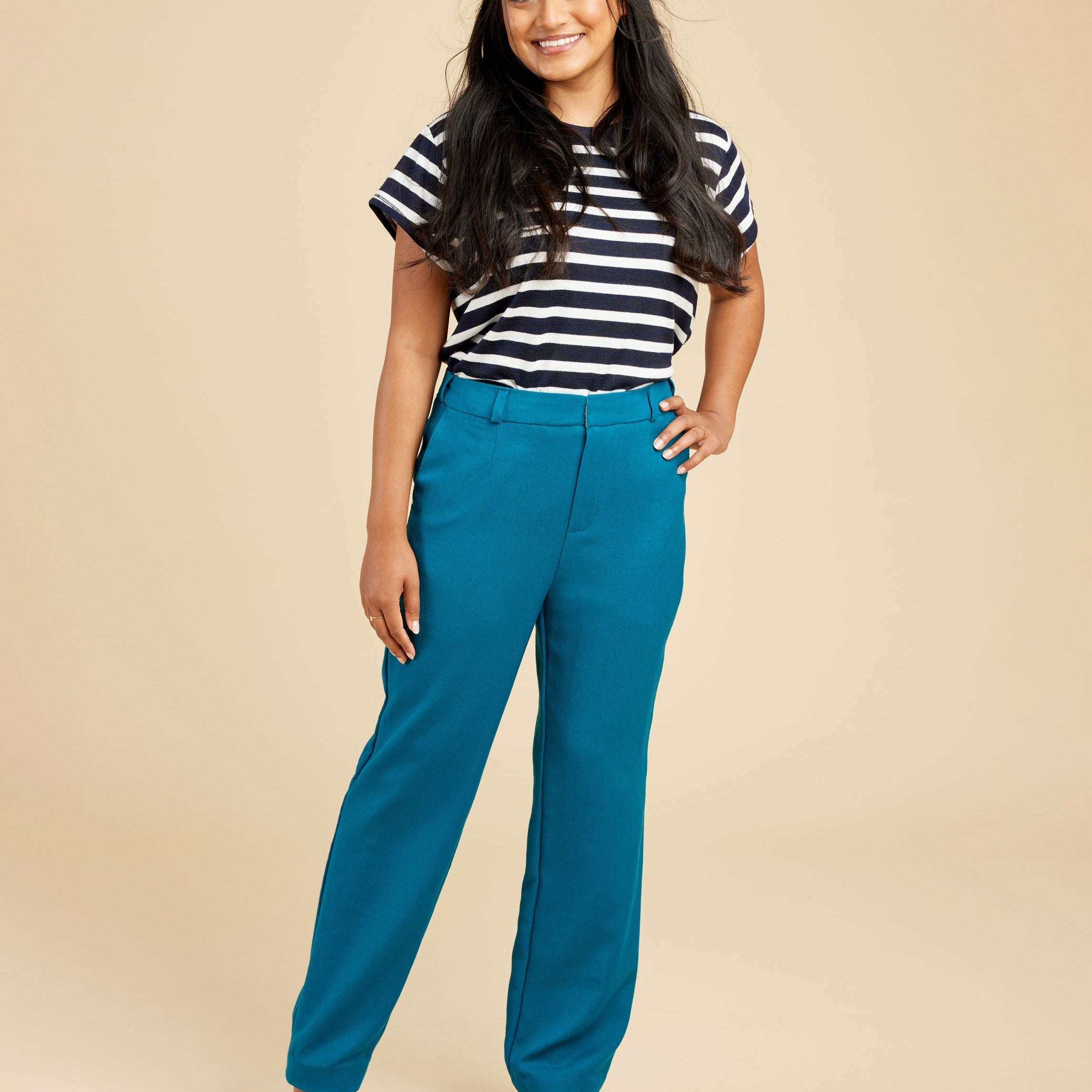 Woman wearing a black and white striped shirt with blue pants on a beige background. These pants are the Meriam Trousers sewing pattern from Cashmerette.