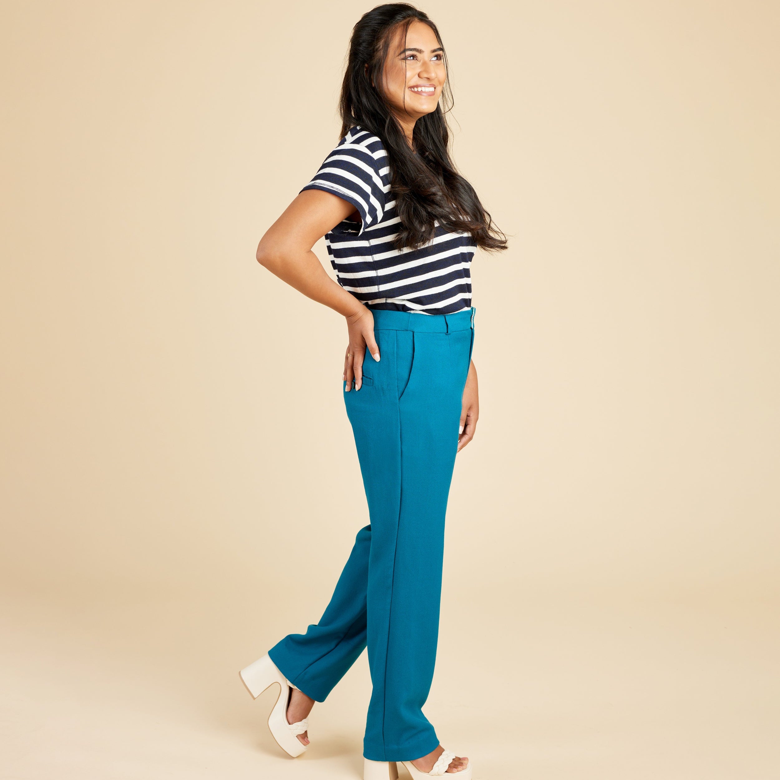 Woman wearing a black and white striped shirt and blue pants on a beige background. These pants are the Meriam Trousers sewing pattern from Cashmerette.
