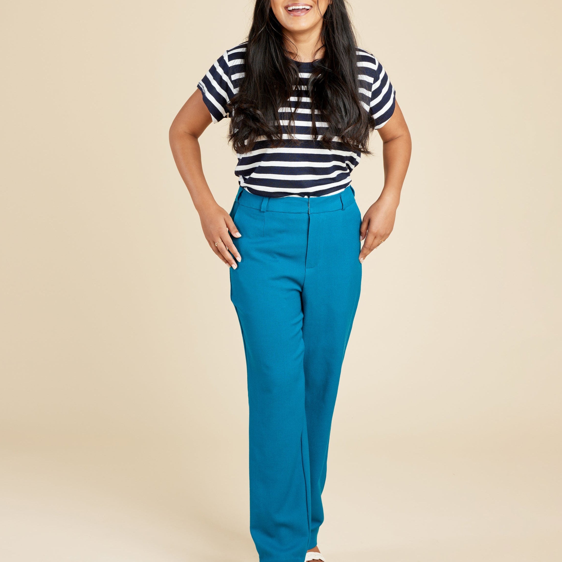 Woman wearing a striped shirt and blue pants on a beige background. These pants are the Meriam Trousers sewing pattern from Cashmerette.