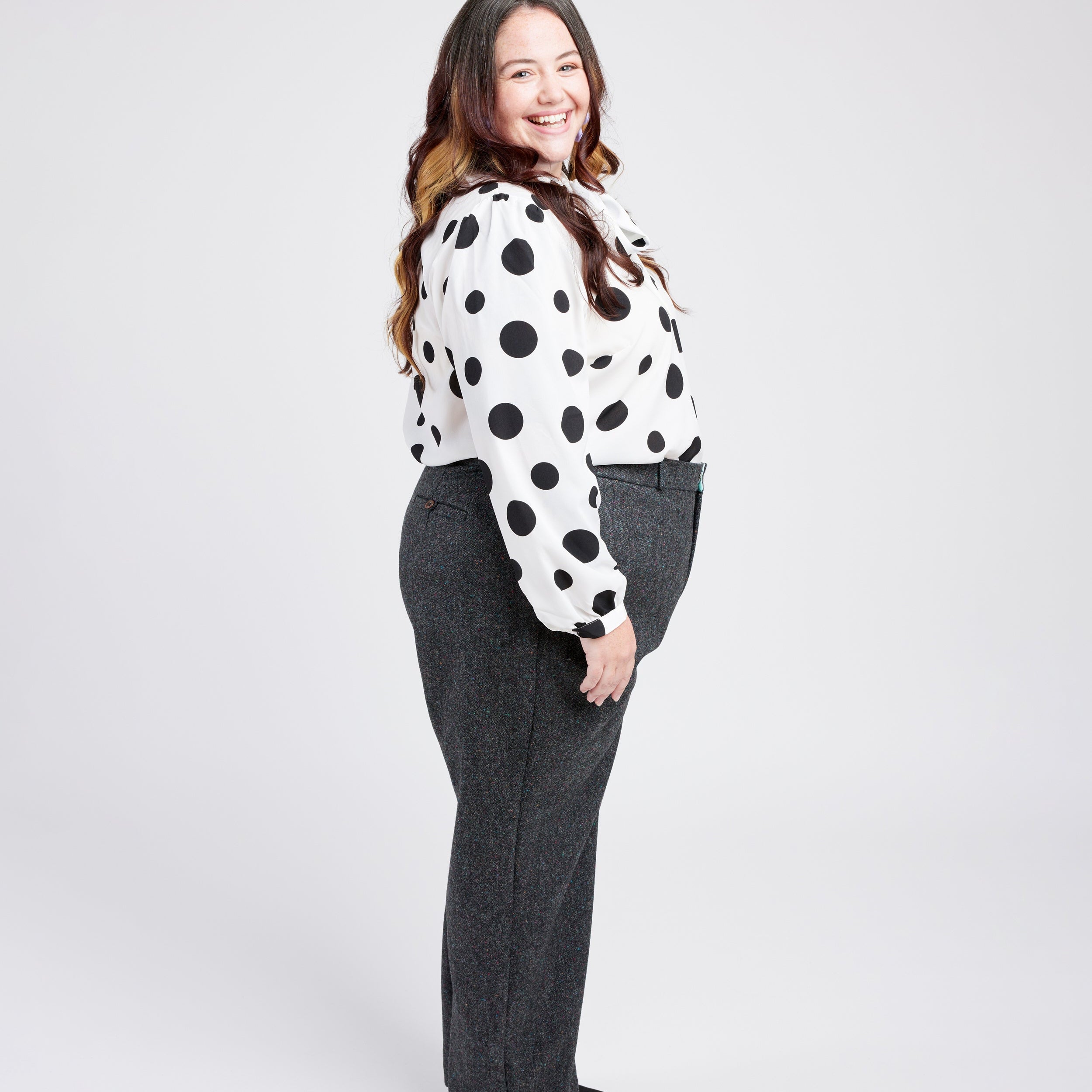 Woman wearing a polka dot blouse and gray pants on a white background. These pants are the Meriam Trousers sewing pattern from Cashmerette.
