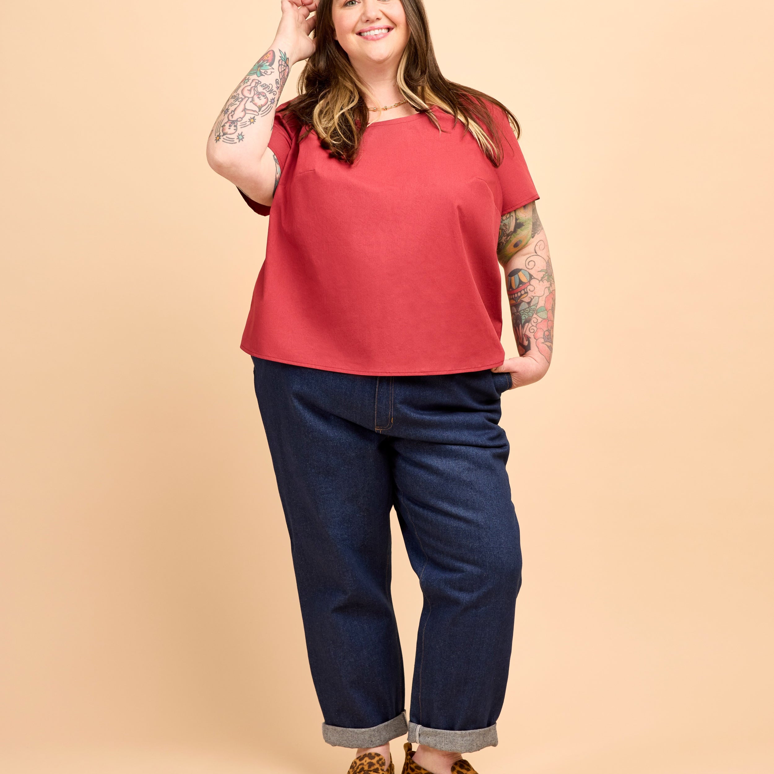 Woman wearing a red top and blue jeans on a beige background. This is the Montrose Top sewing pattern from Cashmerette.