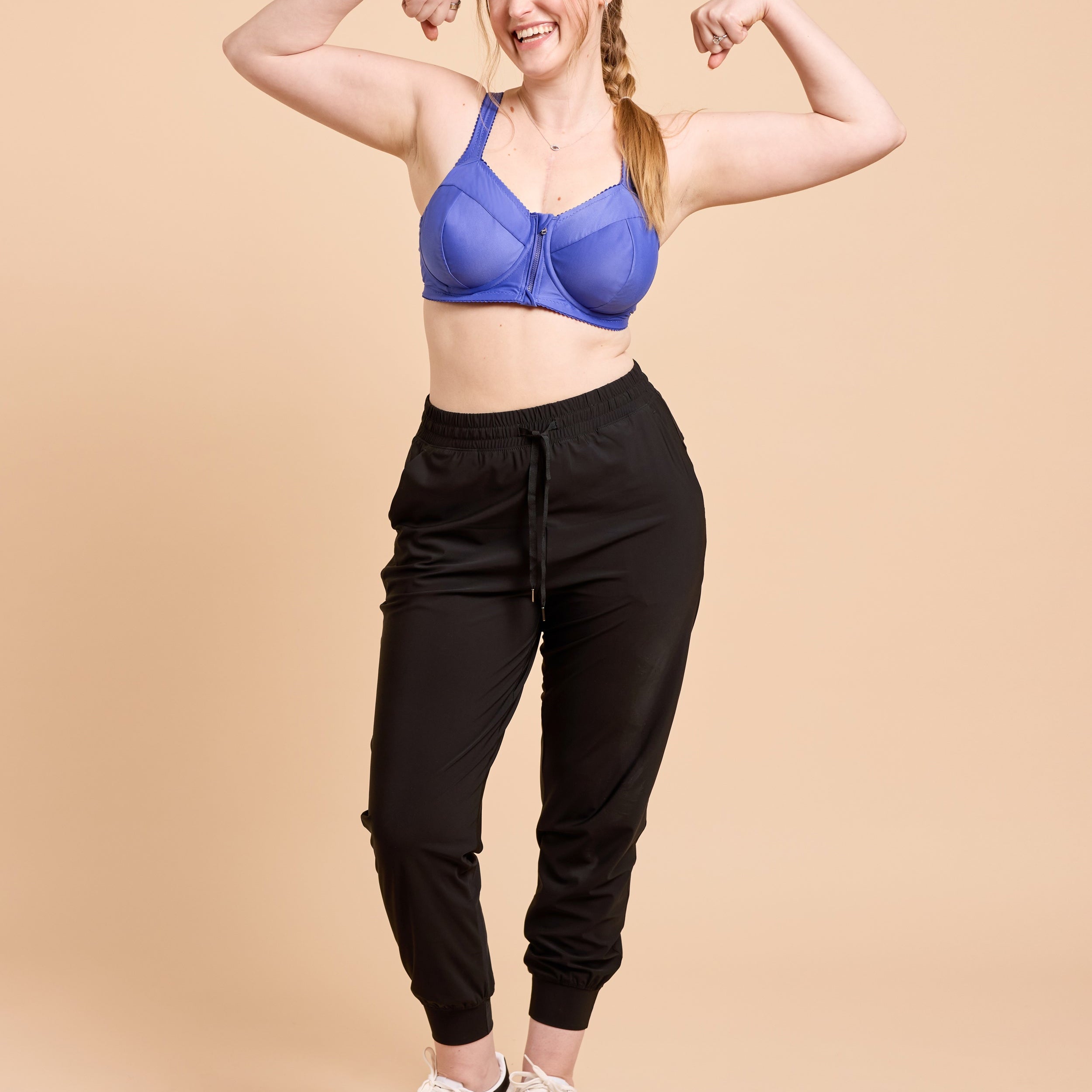 Woman flexing her muscles wearing a blue sports bra and black jogger pants on a beige background. This is the Cashmerette Somerville Sports Bra sewing pattern for big boobs.