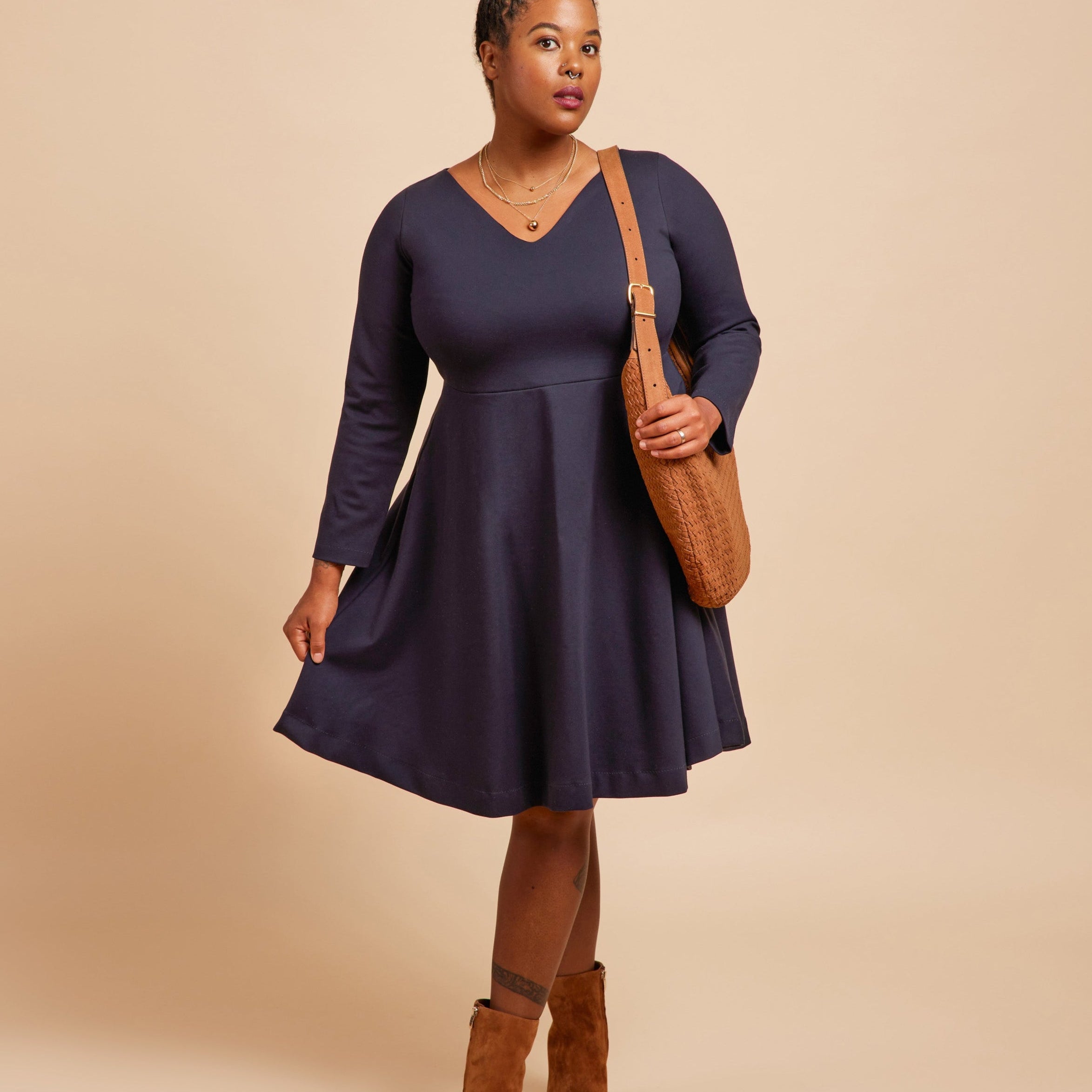 Woman wearing a navy blue dress with brown boots and bag on a beige background. This is the Turner Dress sewing pattern from Cashmerette.