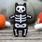 A black felt cat-shaped plush toy with a white skeleton print, standing on a rustic wooden surface with a blurred background of orange and green decorative items.