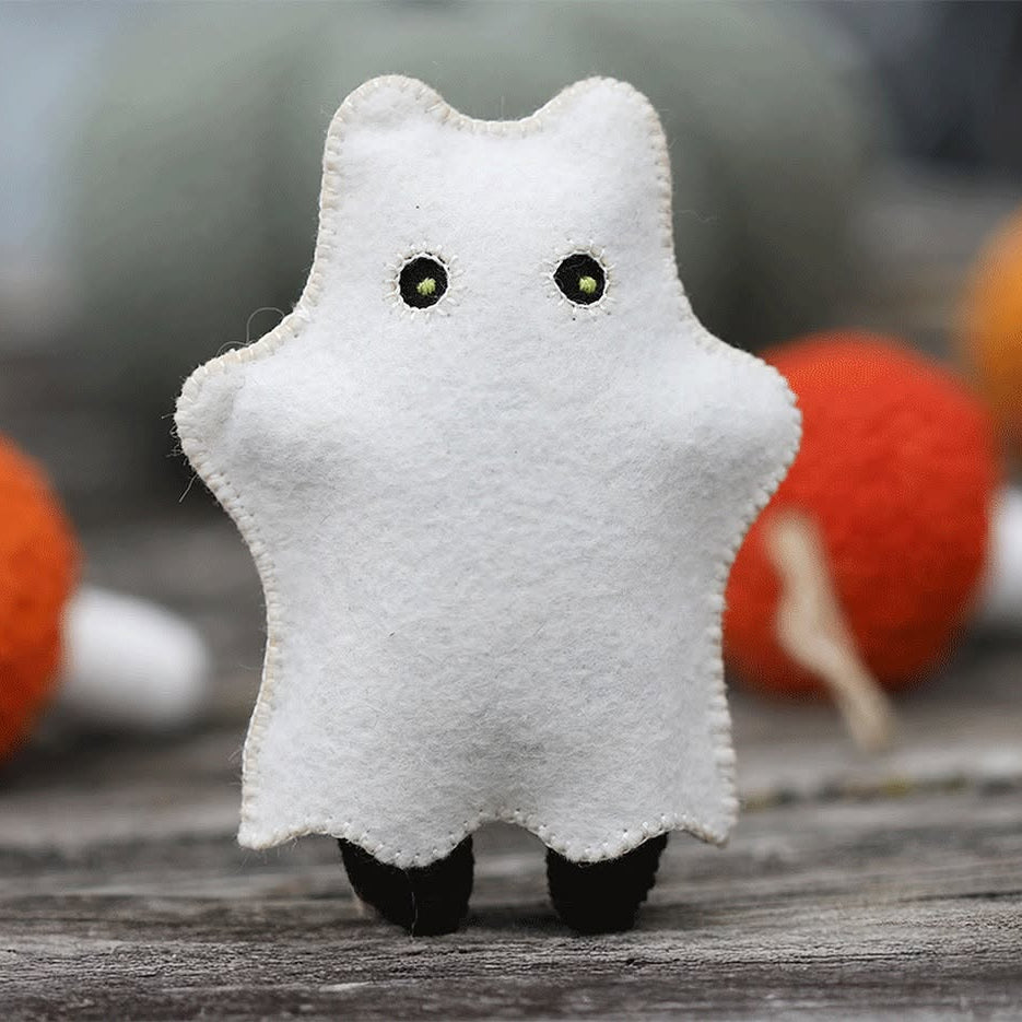 A small white felt ghost plush toy with embroidered eyes and black feet, standing on a rustic wooden surface with blurred orange and gray decorative objects in the background.