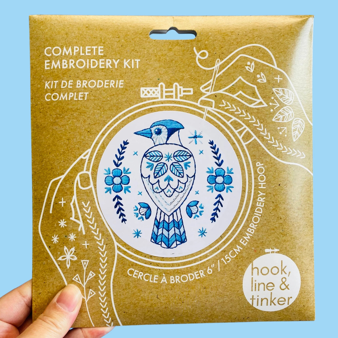 A hand holding a kraft paper package for a complete embroidery kit. The package features a blue bird illustration and white line art of hands holding a needle.