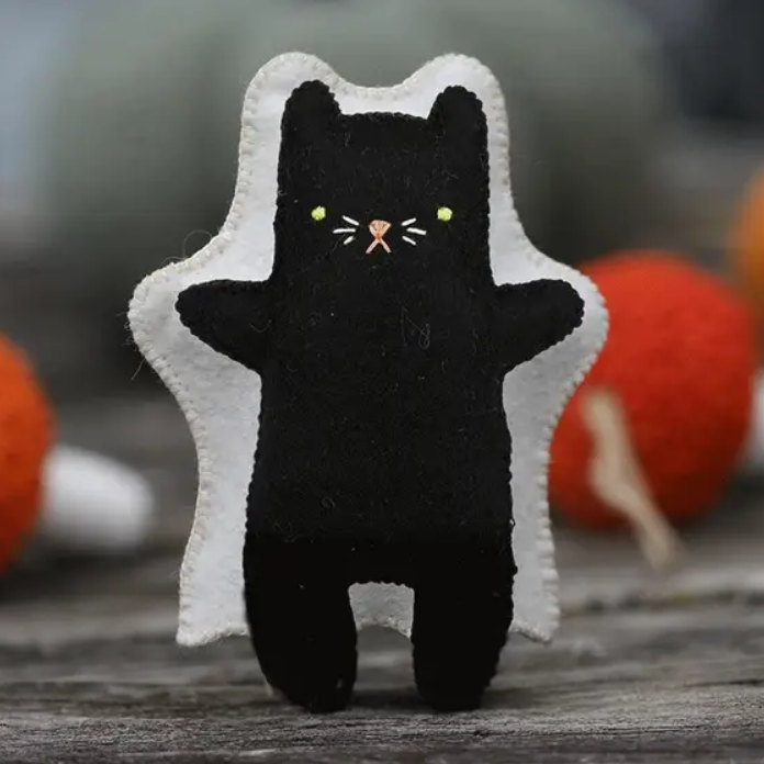 A black felt cat plush toy with a white ghost costume and embroidered face, standing on a rustic wooden surface with blurred orange and red mushrooms in the background.