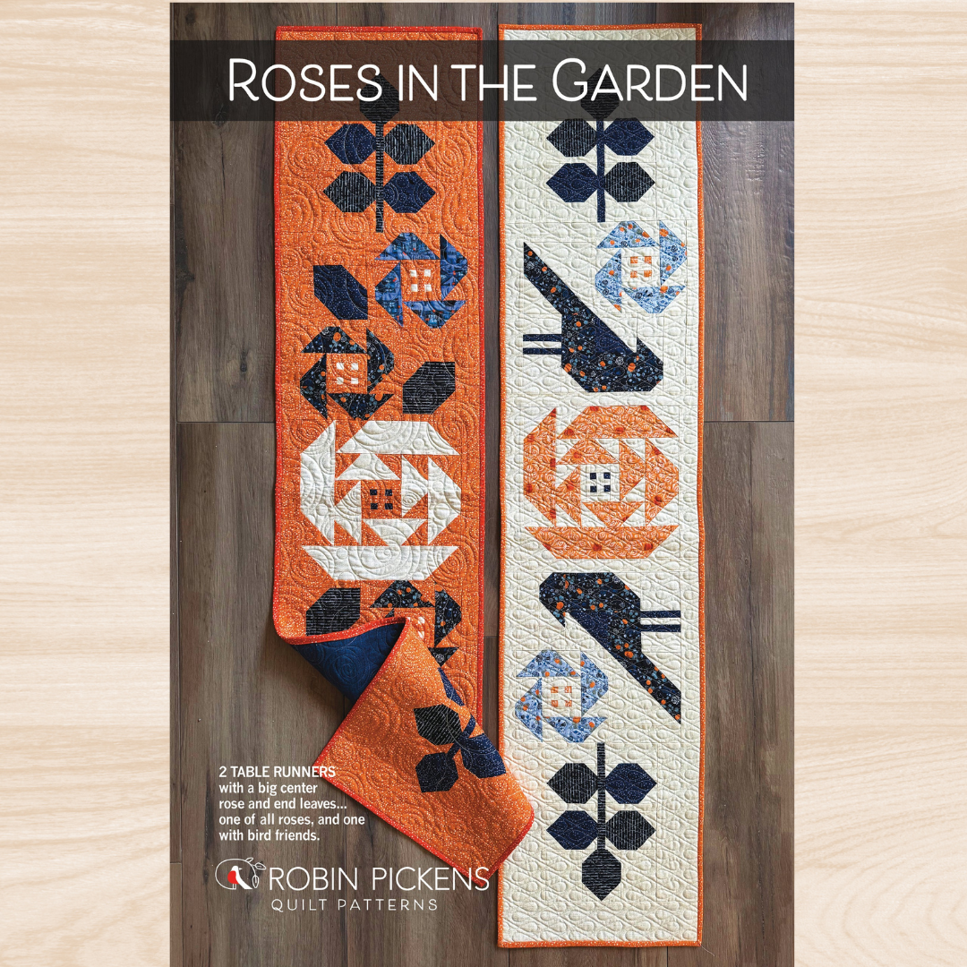 Pattern cover for 'Roses in the Garden' quilt patterns by Robin Pickens, showing two quilted table runners laid flat on a wood surface. The left runner is orange with navy and white floral and bird motifs, and the right runner is off-white with orange and navy floral and bird motifs.