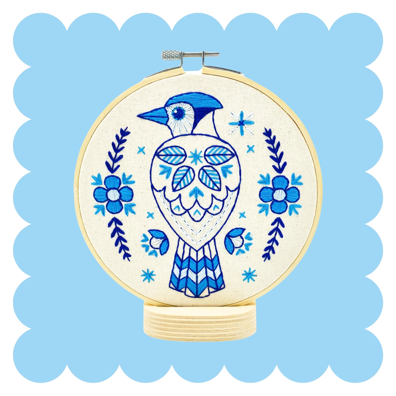 A circular embroidery hoop containing a blue bird and floral design stitched on cream fabric, centered on a light blue scalloped background.