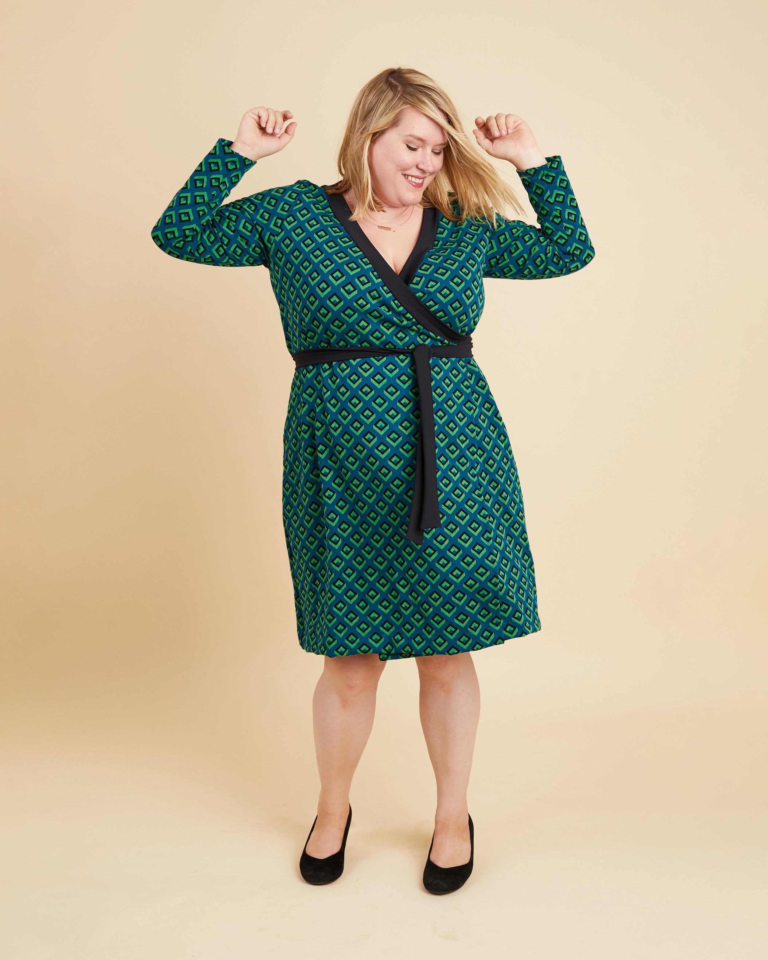 Woman wearing a green patterned dress with a belt on a beige background. This dress is the Appleton Wrap Dress sewing pattern from Cashmerette.