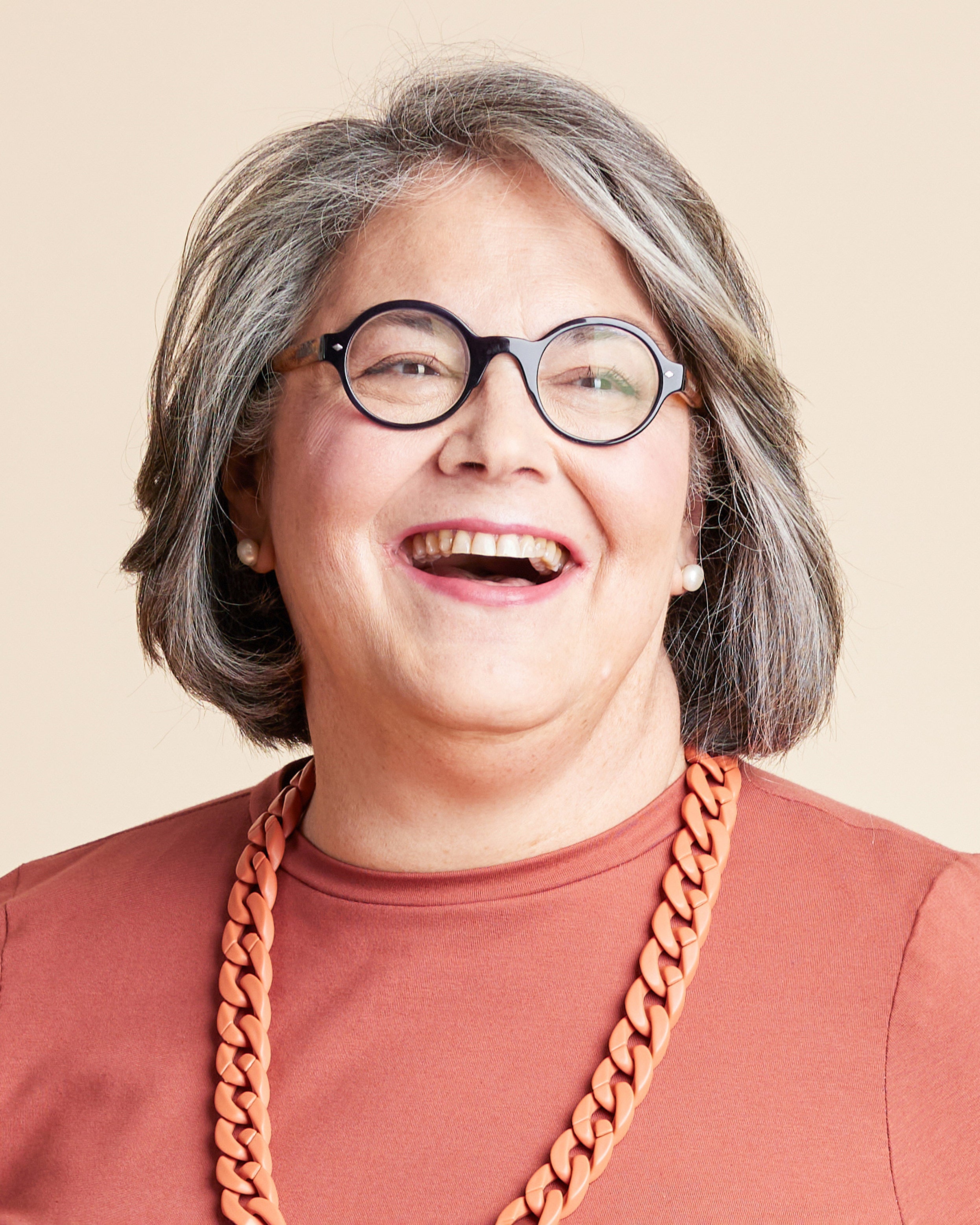 Person with gray hair wearing glasses and a brown shirt with a large orange necklace on a beige background. This is the Grafton Dress, Top & Skirt sewing pattern from Cashmerette.