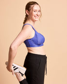 Woman wearing a blue sports bra and black leggings, holding white sneakers against a beige background. This is the Cashmerette Somerville Sports Bra sewing pattern for big boobs.