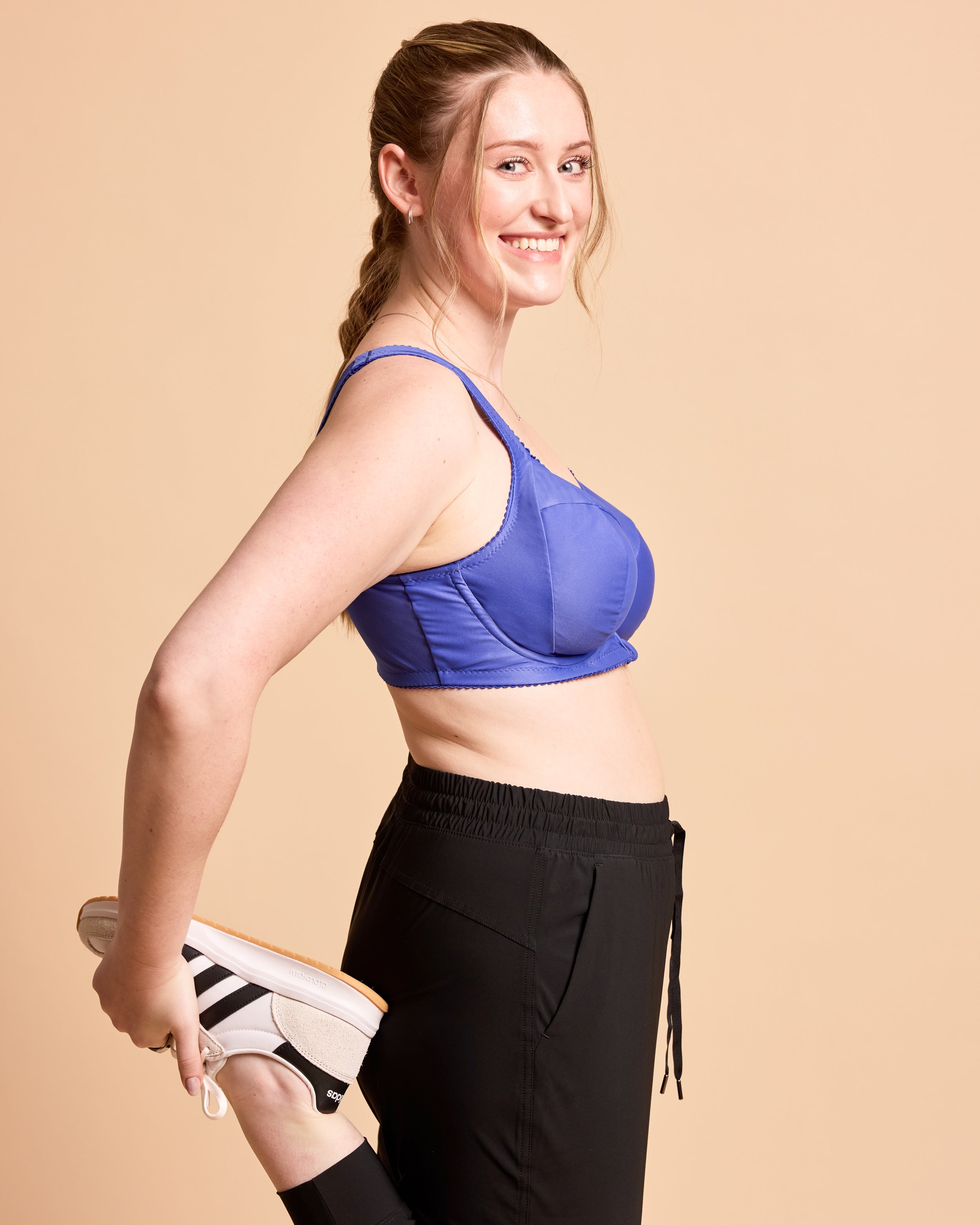 Woman wearing a blue sports bra and black leggings, holding white sneakers against a beige background. This is the Cashmerette Somerville Sports Bra sewing pattern for big boobs.