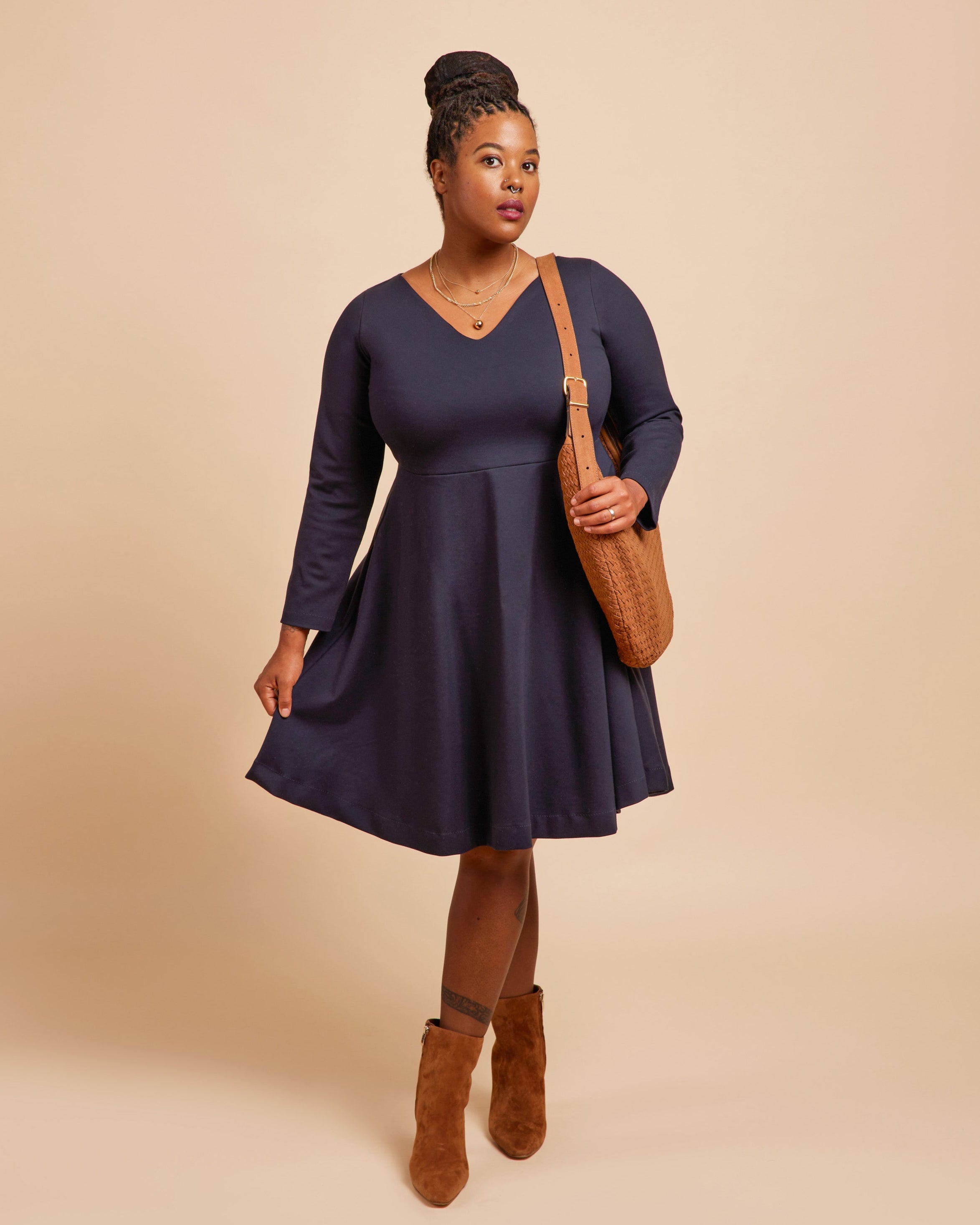Woman wearing a navy blue dress with brown boots and bag on a beige background. This is the Turner Dress sewing pattern from Cashmerette.