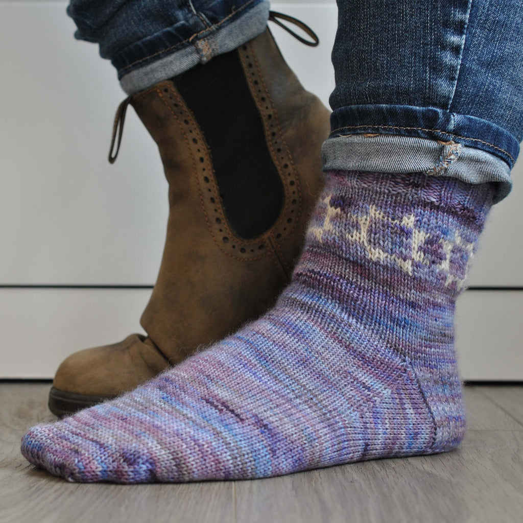 Picture of someone's feet on a wooden floor, one boot is off showing the hand knit sock and the quilt block pattern at its cuff.